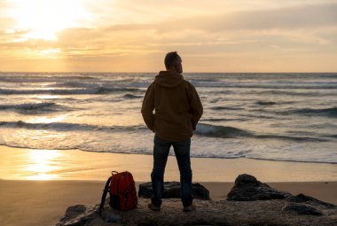 Sırt çantalı sakallı adam gün batımında sıcak güneşli bir günde deniz kenarında tek başına dinleniyor. Seyahat yaşam tarzı konsepti