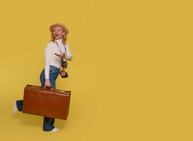 Kot pantolonlu, beyaz gömlekli ve şapkalı bir kadın bavul taşıyor ve sarı arka planda klasik kamerayla fotoğraf çekiyor. Mutlu insanlar tatile çıkıyor.