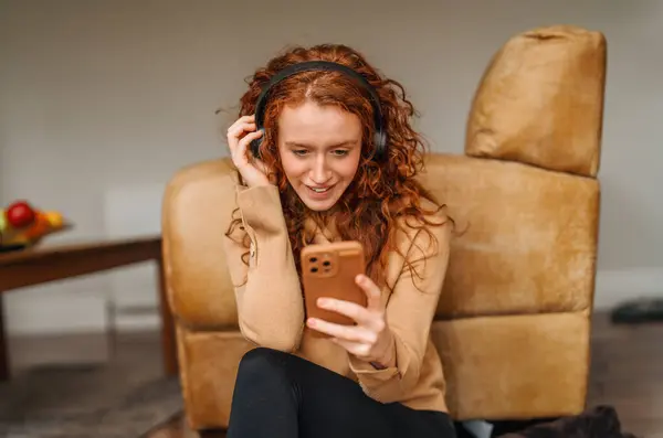 Happy young redhead woman in brown shirt and using a mobile phone, listening to favorite songs, audiobooks, podcasts on headphones while sitting near a couch at home
