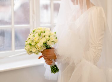 Pencerenin yanında şık bir düğün saç stili olan güzel bir gelin. Yakından, elinde bir gelin buketi tutan muhteşem gelinin portresi. Düğün için. Yüzünde peçeyle gelin portresi