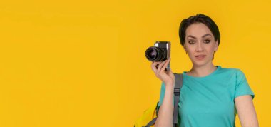 Gülümseyen kadın fotoğrafçı, sırt çantalı bir seyahat blogcusu fotoğraf makinesi, stüdyo fotoğrafı, sarı arka plan..