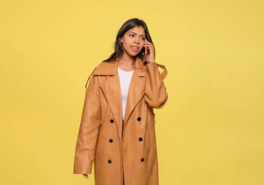 Kot pantolonlu bir kadın, beyaz bir tişört ve sarı arka planda oturan cep telefonu kullanan kahverengi bir ceket. Mutlu insanlar.
