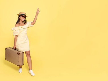 Sarı elbiseli ve şapkalı bir kadın, bavul taşıyor ve sarı arka planda klasik kamerayla fotoğraf çekiyor. Mutlu insanlar tatile çıkıyor.
