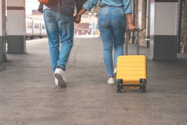 Bavulları ve sırt çantaları ile mutlu bir çift tren yolu platformu boyunca yürüyorlar ve tatile çıkmaya hazırlar. Erkek ve kadın el ele tutuşuyor, geriye bakıyor ve gülümsüyor. Tren konseptiyle seyahat