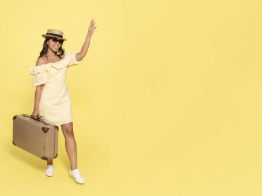 Sarı elbiseli ve şapkalı bir kadın, bavul taşıyor ve sarı arka planda klasik kamerayla fotoğraf çekiyor. Mutlu insanlar tatile çıkıyor.
