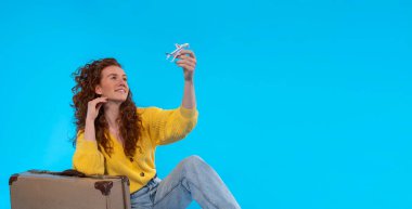 Sarı ceketli, elinde bavulu ve oyuncak uçağıyla oturan ve mavi arka planda bir yolculuğun hayalini kuran mutlu, kızıl saçlı kadın. Seyahat kavramı