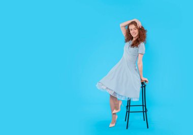 Mavi elbiseli kızıl saçlı harika bir kadın mavi arka plandaki taburede oturuyor.