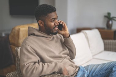 Kahverengi kapüşonlu mutlu adam telefon kullanıyor, çalışıyor, öğreniyor, alışveriş yapıyor, internette çalışıyor, evde bir kanepede otururken konuşuyor.