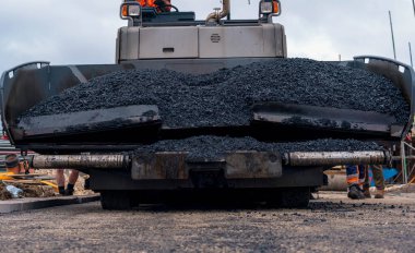 Asphalt paver filled with hot tarmac laying new road surface on new residential housing development site and roadworker operator in orange hi-viz next to it clipart