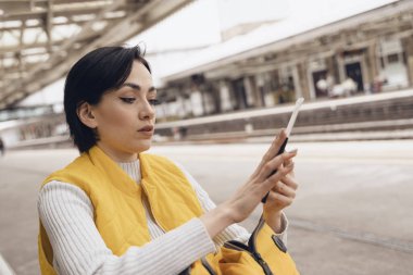 Sarı ceketli genç esmer kadın elinde pasaport ve biletlerle tren istasyonunda tren bekliyor. Seyahat kavramının tadını çıkarıyorlar.