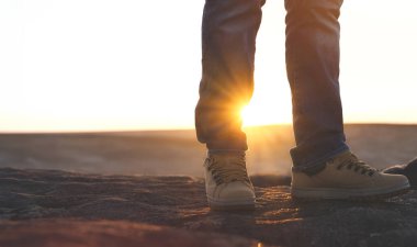 Özel botlu bir adamın ayakları dağlarda yürürken ve gün doğumunda ya da günbatımında dağın tepesine çıkarken. Seyahat yaşam tarzı konsepti