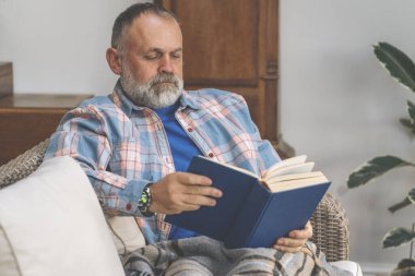 Yaşlı, yaşlı bir adam gülüyor kitap okuyor, dinleniyor, oturma odasında kanepede oturuyor.