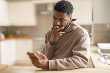 Kahverengi kapüşonlu düşünceli bir adam telefon kullanıyor, çalışıyor, öğreniyor, alışveriş yapıyor, internette çalışıyor, evde bir kanepede otururken konuşuyor.