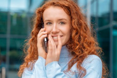 Beyaz tişörtlü, cep telefonuyla konuşan ve kullanan modern bir şehirde cep telefonu kullanan genç bir kızıl saçlı. Yaşam tarzı fotoğrafları