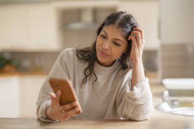 Gri gömlekli mutlu bir Hintli kadın cep telefonu kullanıyor, favori şarkıları, müziği, sesli kitapları, podcast 'leri dinliyor, mutfakta otururken konuşuyor.