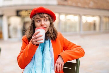 Kırmızı paltolu ve bereli genç bir kadın dışarıdaki kafede kahve içiyor, telefonda konuşuyor ve eğleniyor. yaşam tarzı konsepti