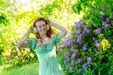 Açık havada, yeşil elbiseli ve kulaklıklı, halka açık parkta dans eden mutlu kızıl saçlı kadının portresi. Çevrimiçi müzik dinleyip kendin ol.