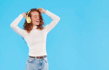 Mutlu, genç, kızıl saçlı bir kadın, müzikle, tasasız kollarıyla kulaklarında stereo kulaklıklarla, mavi arka planda beyaz gömlek ve pantolonla uzaklara taşınıyor.