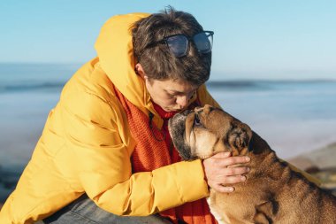 Sarı ceketli olgun bir kadın köpekle dağlarda yürüyüş yapıyor, sağlık için egzersiz ve uygunluk, sağlıklı yaşam tarzı ve gülümseme. Olgun bir dişi buldogla bir kayanın üzerinde oturuyor, sakin bir gün.