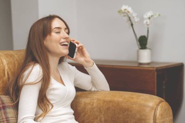 heyecanlı kızıl saçlı kadın koltukta oturuyor, evdeki oturma odasında cep telefonu kullanıyor.