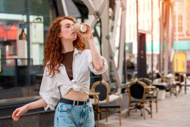 Kot pantolon ve beyaz tişörtlü mutlu, genç, kızıl bir kadın kahve içiyor ve eğleniyor. yaşam tarzı konsepti