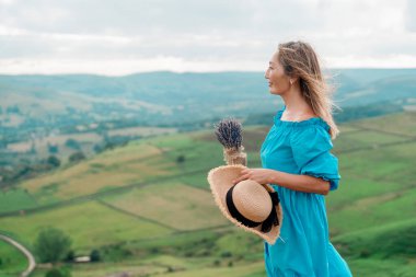 Yaz günbatımında dağın tepesinde lavanta desenli mavi elbiseli ve şapkalı Asyalı bir kadın.