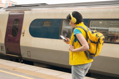 Kulaklıklı kadın cep telefonundan tren tarifesini kontrol ediyor.