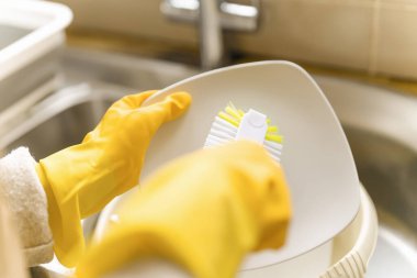 Sarı eldivenler giymiş, mutfak lavabosunda fırçayla beyaz tabak fırçalayan iki el.