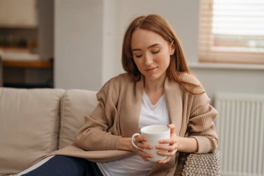 Bir kadın kanepede otururken sıcak bir içecekten hoşlanır..