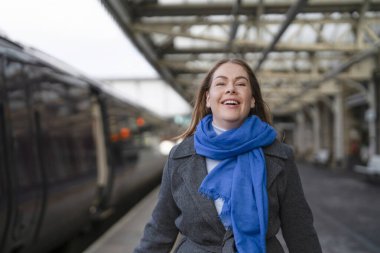 Mavi eşarplı bir kadın tren istasyonuna vardığında parlak bir şekilde gülümsüyor..