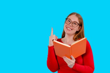 Kırmızı gömlekli ve gözlüklü gülümseyen bir kadın turuncu bir kitap tutuyor ve işaret parmağıyla yukarı doğru işaret ediyor..