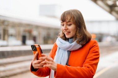 Turuncu ceketli ve mavi eşarplı gülümseyen genç kadın tren istasyonunda akıllı telefon kullanırken gülümsüyor..
