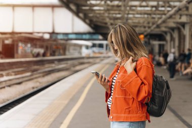 Parlak turuncu ceketli kadın tren istasyonunda bekliyor, telefona bakıyor..