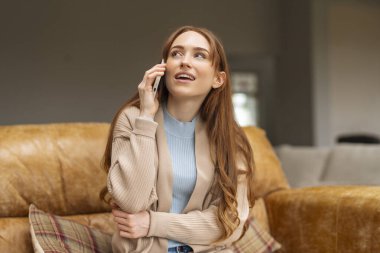 Uzun kızıl saçlı mutlu genç bir kadın koltukta oturuyor cep telefonuyla konuşuyor mavi gömleğin üzerine bej bir hırka giyiyor.