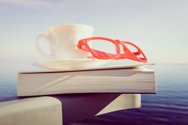 Tatillerde kitap okumak. Kahve fincanı ve kitap açık havada mavi deniz suyuna karşı sandalyede..