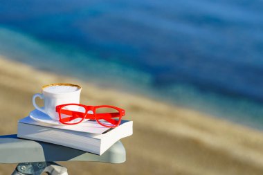 Ruhsal iyileşme, ruhsal sessizlik. Sahile karşı kahve fincanı, kitap ve kırmızı bardaklar. Tatilde okumak. Tatillerde dinlenmek. Zihinsel sağlık sorunları.