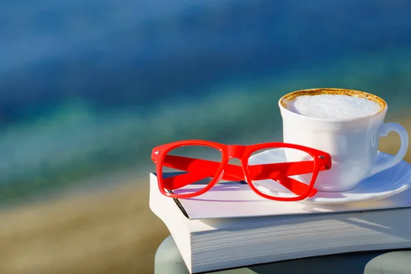 Spiritual healing, psyche silence. Coffee cup, book and red glasses against coast. Reading on vacation. Relaxation on holidays. Mental health break.