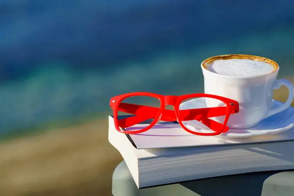 Spiritual healing, psyche silence. Coffee cup, book and red glasses against coast. Reading on vacation. Relaxation on holidays. Mental health break.