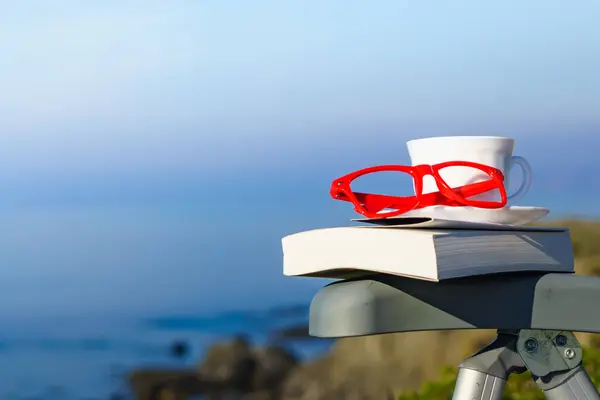 Spiritual healing, psyche silence. Coffee cup, book and red glasses against coast. Reading on vacation. Relaxation on holidays. Mental health break.