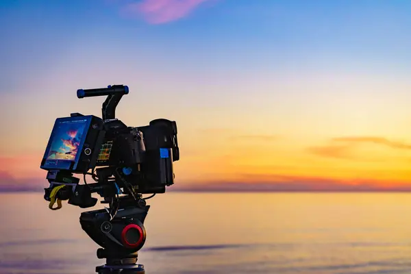 Professional camera on tripod taking picture film video of sunset over sea.
