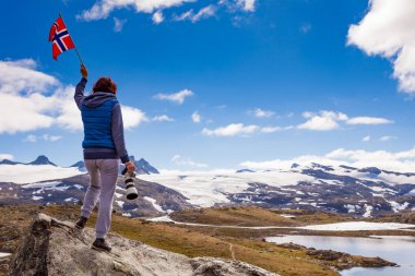 Tourist woman enjoy mountains landscape, holding norwegian flag and photo camera. National tourist scenic route 55 Sognefjellet, Norway clipart