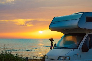 Akdeniz kıyısındaki karavan ve deniz üzerinde gün doğumundan fotoğraf çeken üç ayaklı kamera. Doğa plajında kamp yapmak. Karavanla tatil gezisi.