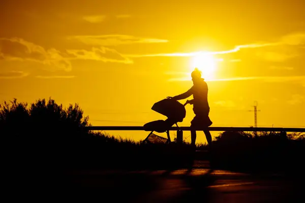 日落时分带着婴儿车在海滨散步的妇女的轮廓 图库照片