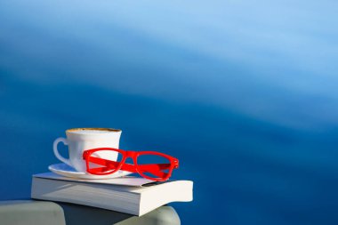 Tatilde okumak. Kahve fincanı, kitap ve kırmızı bardaklar mavi deniz suyuna karşı. Tatil gezisinde dinlenmek. Zihinsel sağlık sorunları.