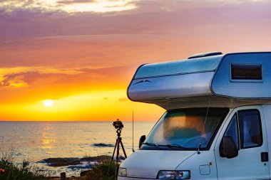 Akdeniz kıyısındaki karavan ve deniz üzerinde gün doğumundan fotoğraf çeken üç ayaklı kamera. Doğa plajında kamp yapmak. Karavanla tatil gezisi.