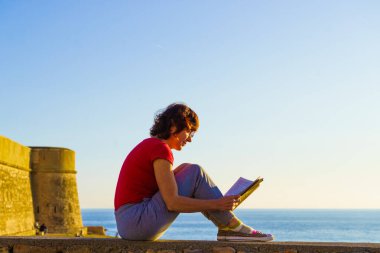 Olgun turist kadın kitapla sahilde dinleniyor, temiz havanın tadını çıkarıyor. Tatillerde kitap okumak. Dinlenme zamanı..