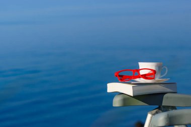 Tatilde okumak. Kahve fincanı, kitap ve kırmızı bardaklar mavi deniz suyuna karşı. Tatil gezisinde dinlenmek. Zihinsel sağlık sorunları.