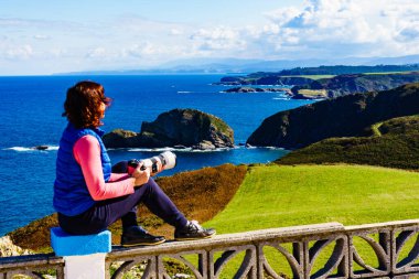 Woman with camera take travel photo from seascape of Atlantic ocean and Asturias coast at Cape Penas in north Spain. clipart