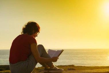 Olgun bir turist kadın kitapla sahilde dinleniyor, gün batımında temiz havanın tadını çıkarıyor. Tatillerde kitap okumak. Dinlenme zamanı..