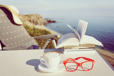 Tatillerde kitap okumak. Kahve fincanı ve kitaplar açık havada mavi deniz suyuna karşı. Kitap sayfaları rüzgarda çırpınıyor.
